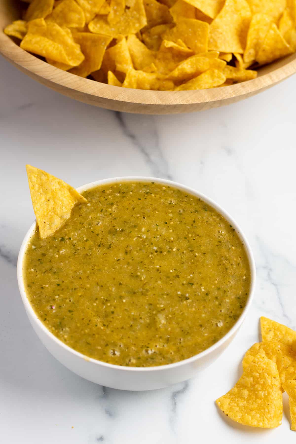 A white bowl with a green tomatillo salsa and a wooden bowl with tortilla chips. 