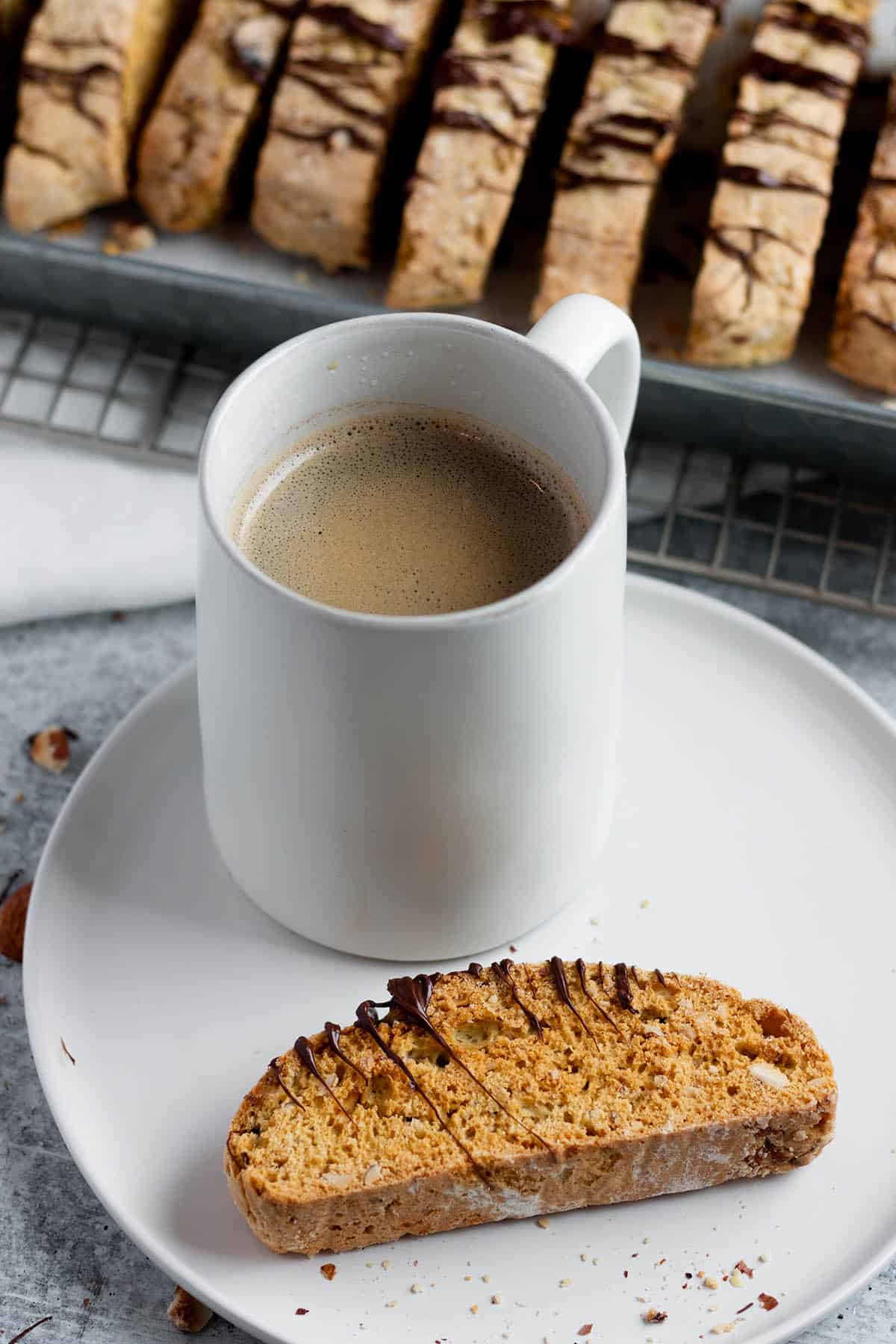 A plate with a piece of almond biscotti, a mug of coffee and a metal rack with more biscotti in the back. 