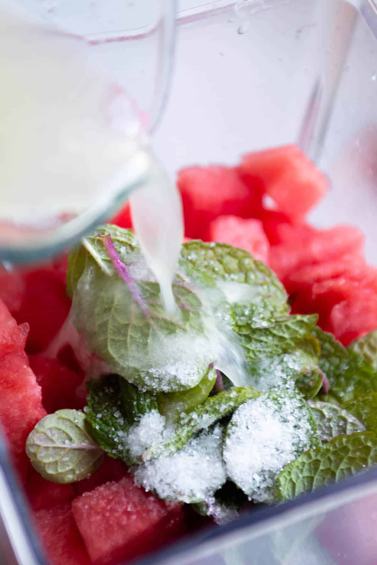 Blender jar with diced watermelon, mint leaves, salt and lime juice being poured in. 