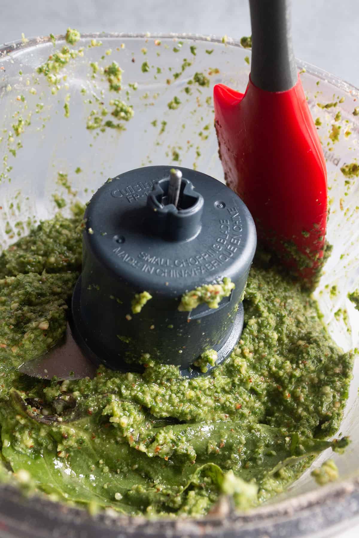A spatula scraping the sides of the food processor to incorporate pieces that didn't puree back into the pesto sauce. 