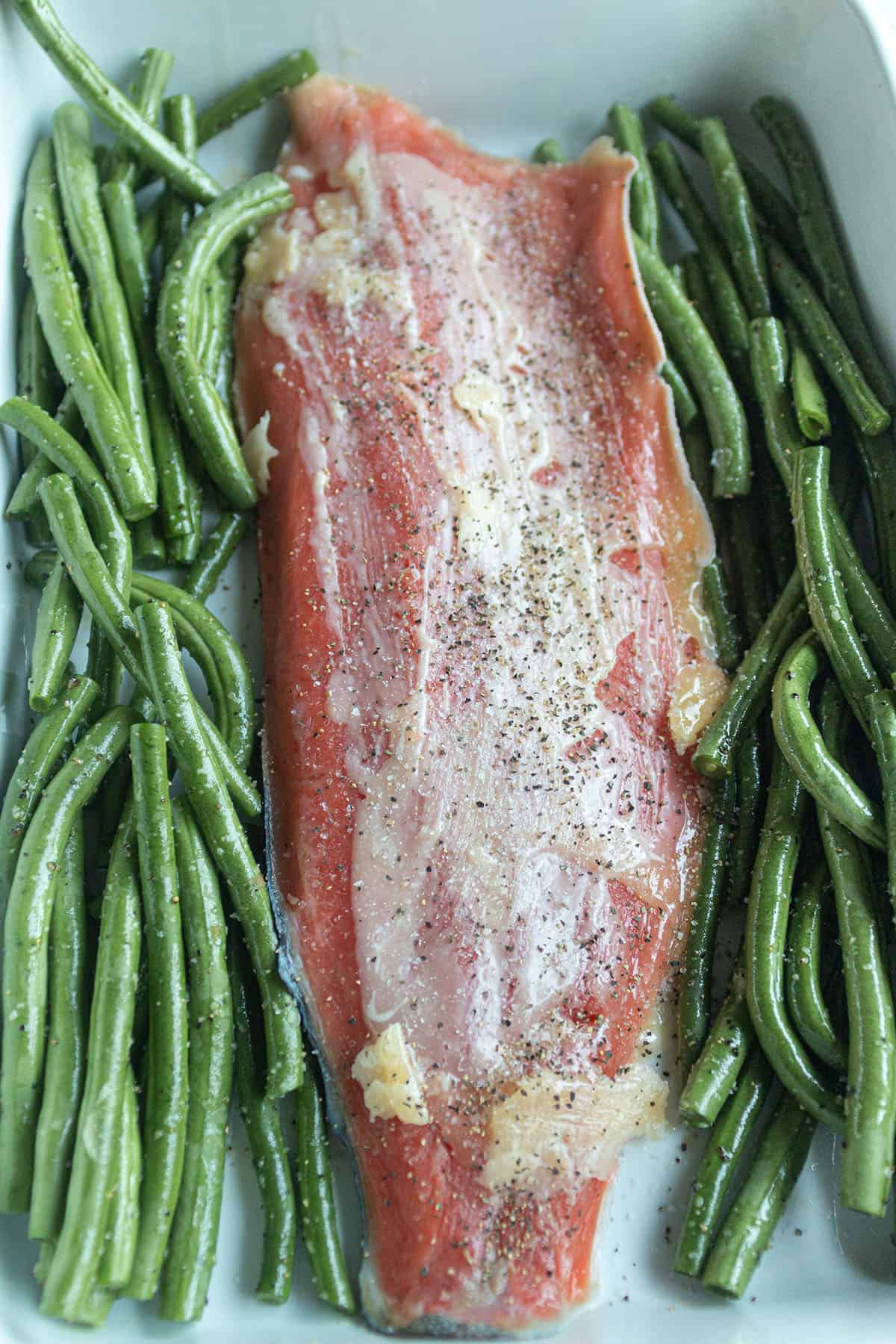 Baking dish with a salmon fillet covered with butter topped with salt and pepper with a side of green beans.