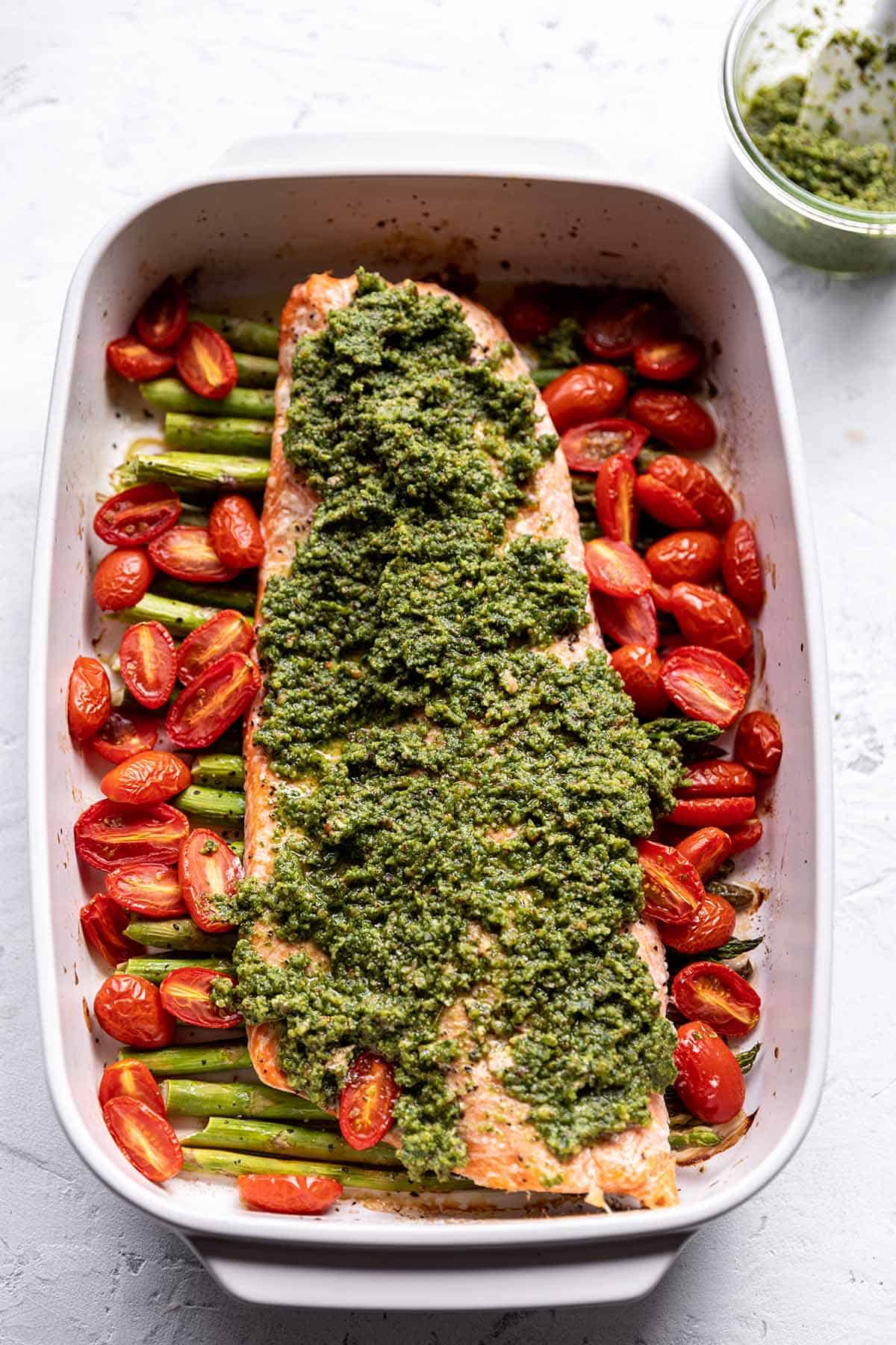 A baking pan with asparagus, cherry tomatoes, a salmon filet and pesto on top.
