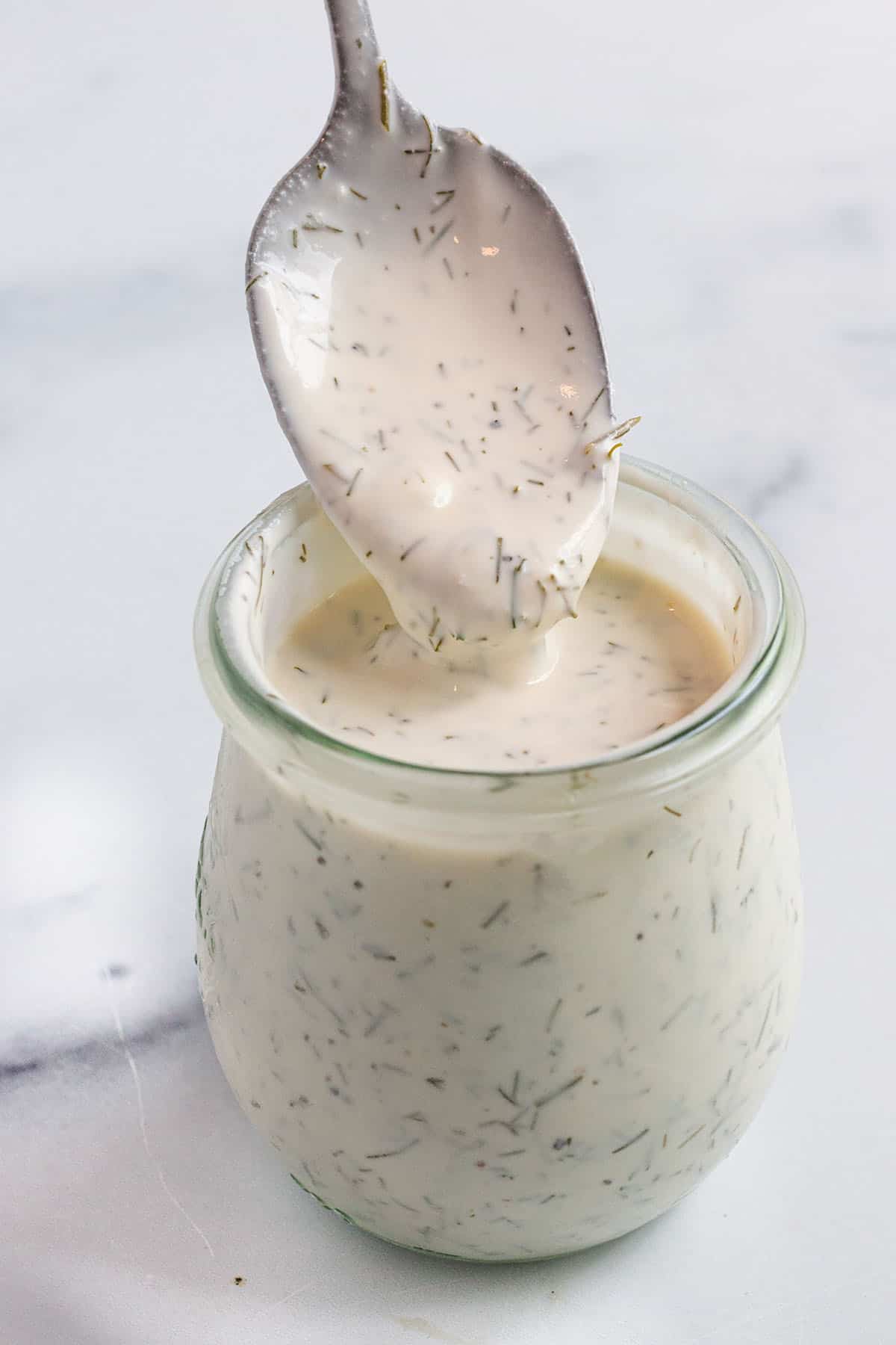 A small weck jar with a creamy white dill sauce and spoon scooping some out. 