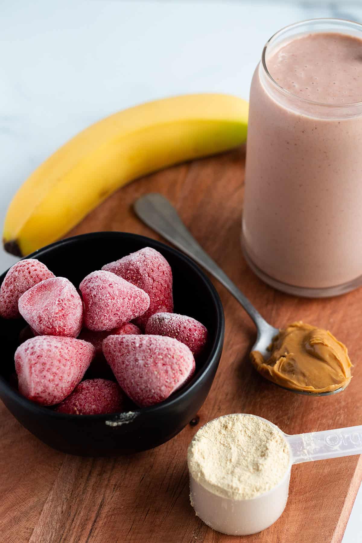 A wooden board with a smoothie, banana, frozen strawberries, protein powder and peanut butter.