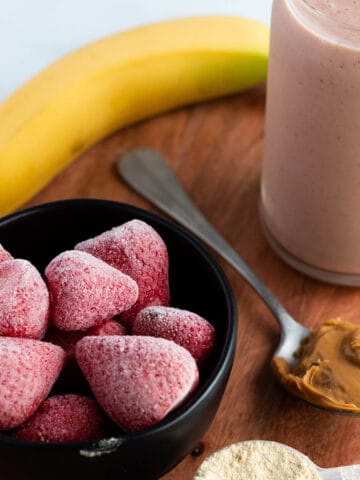 A wooden board with a glass filled with a pink smoothie, banana, frozen strawberries, scoop of peanut butter and protein powder.