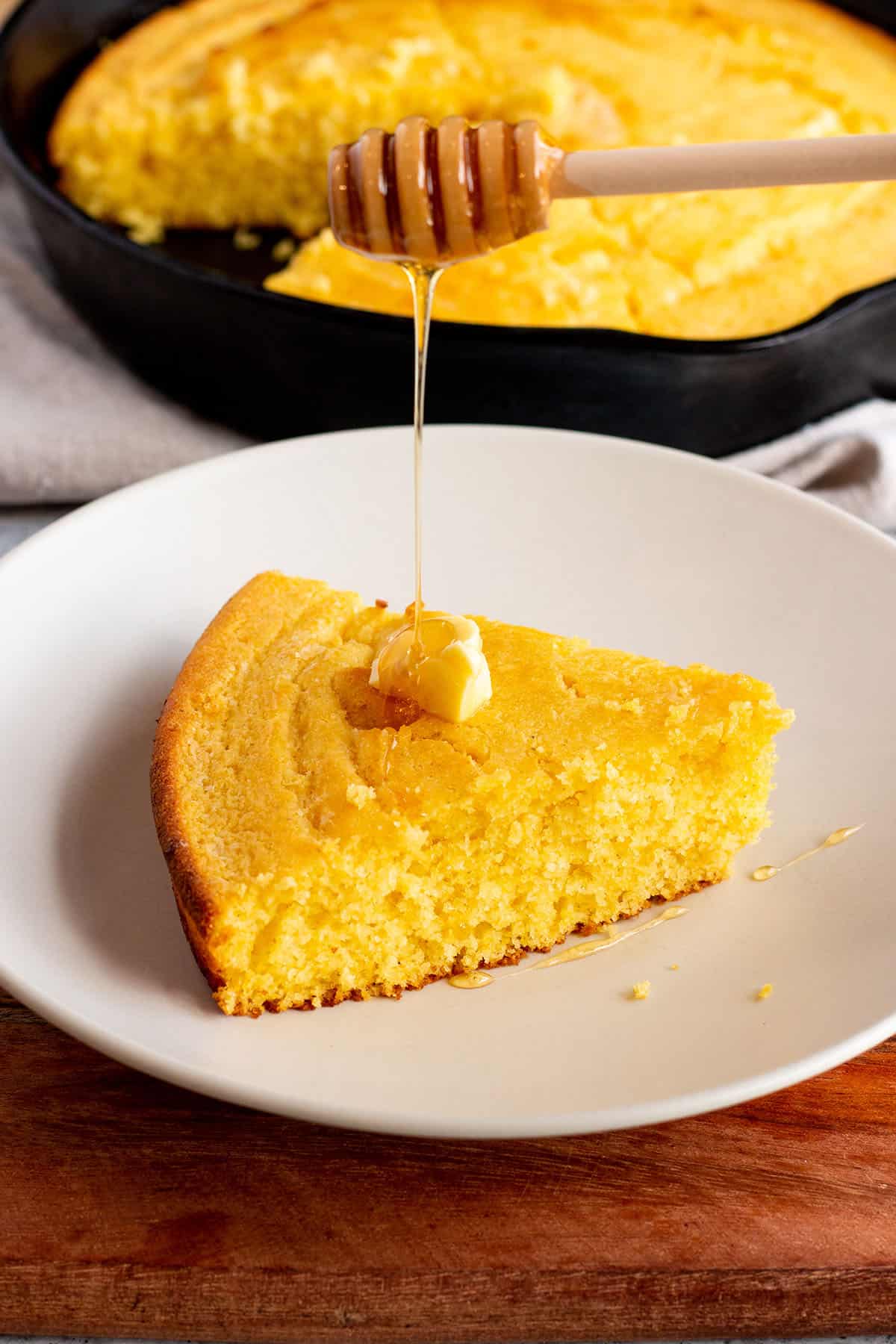 A wedge of cornbread with honey being drizzled on with a cast iron pan in the back. 