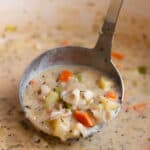 A ladle scooping out some clam chowder with bits of clam, potatoes, carrots and celery.