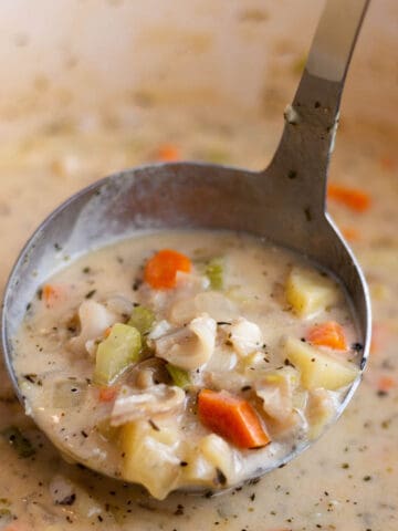 A ladle scooping out some clam chowder with bits of clam, potatoes, carrots and celery.