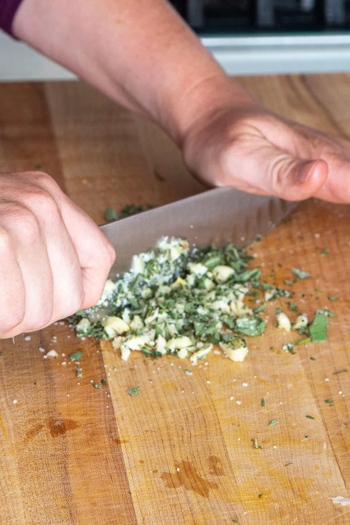 Two hands with a chefs knife chopping herbs, garlic and salt together. 