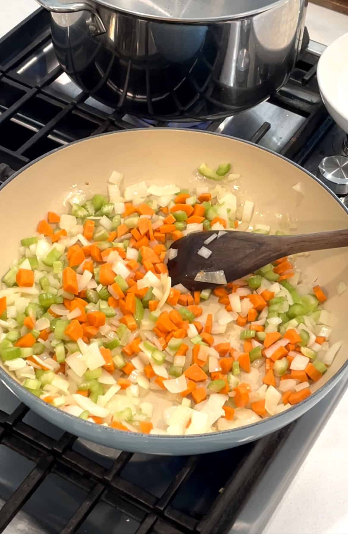 Large skillet with sautéed onions, carrots, and celery with a wooden spatula. 