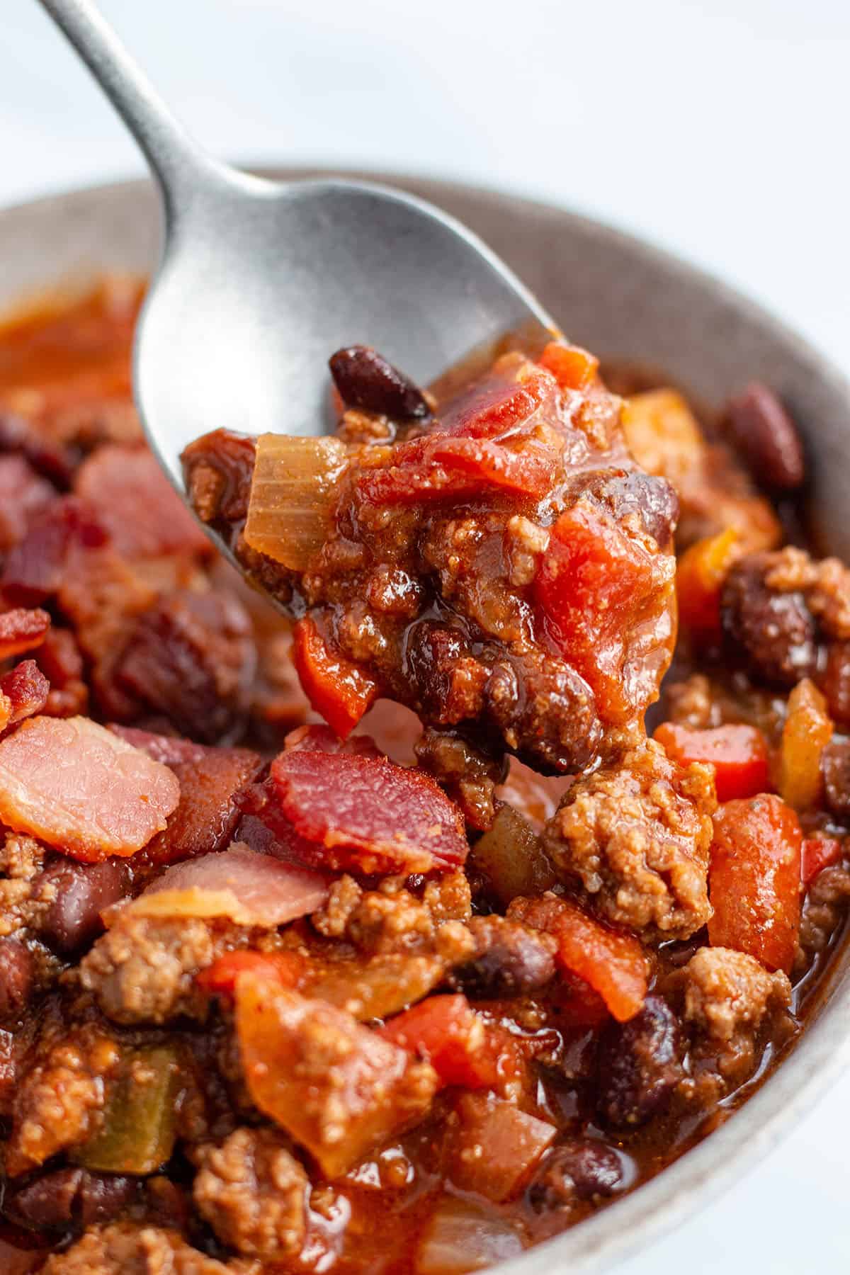A bowl full of chili with beef, black beans, peppers, and bacon with a spoon scooping some out. 