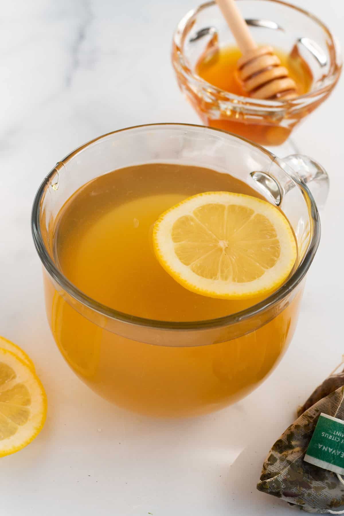 A clear coffee mug with a medicine ball tea inside and fresh lemon with tea bags and a bowl of honey in the back.