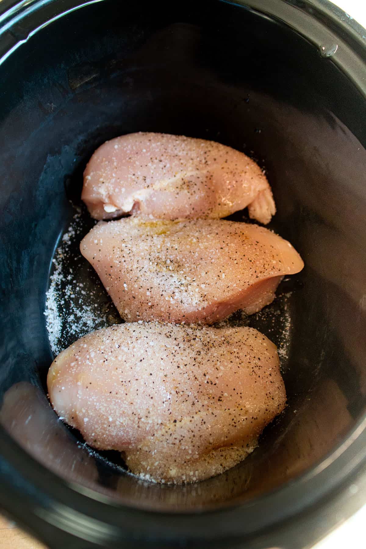 A black crockpot with raw chicken with salt and pepper.