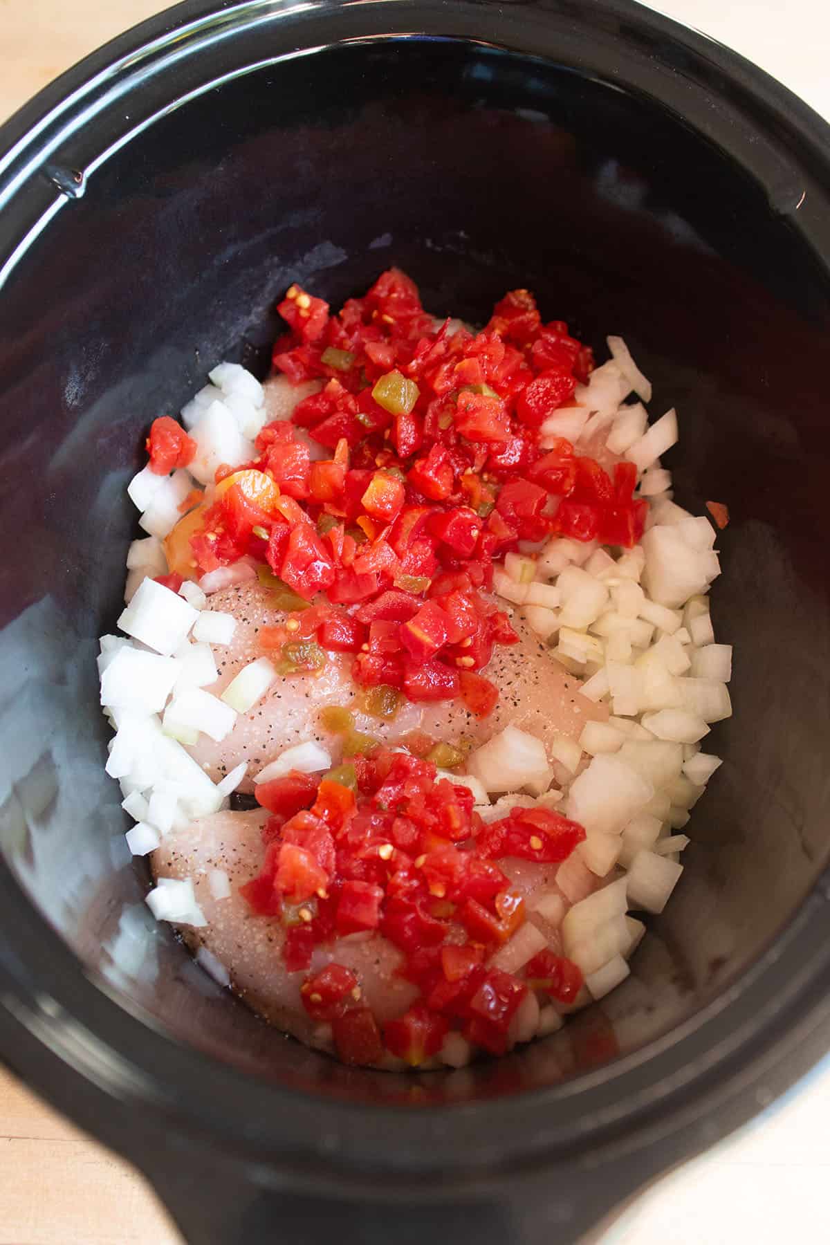 A slow cooker with raw chicken, onions and diced tomatoes.