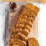 A wooden board on a cooling rack with a sliced loaf of pumpkin bread with a jar of cinnamon sticks.