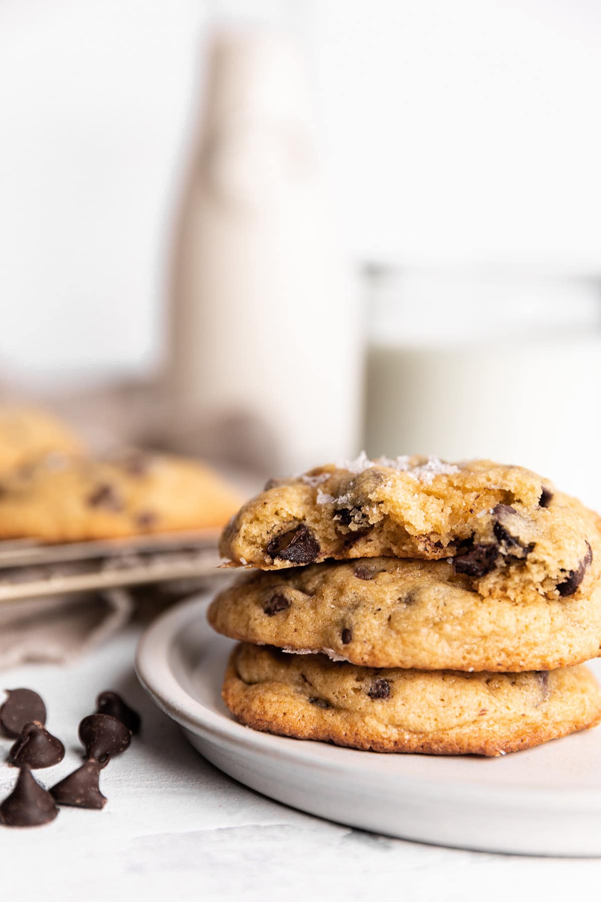 A stack of chocolate chip cookies on a plate with a glass and container of milk in the back. 