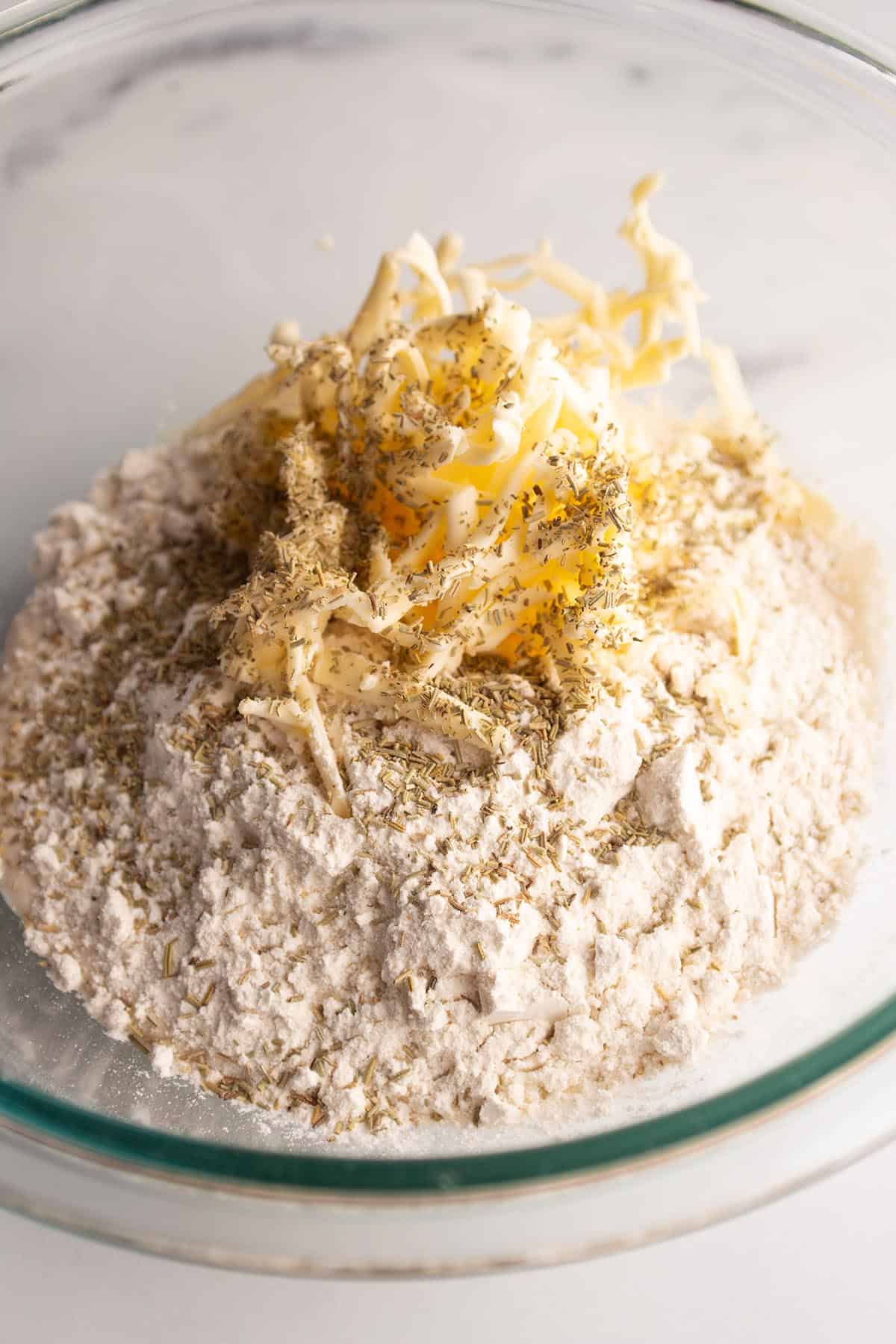 A glass bowl with flour, grated butter and rosemary.