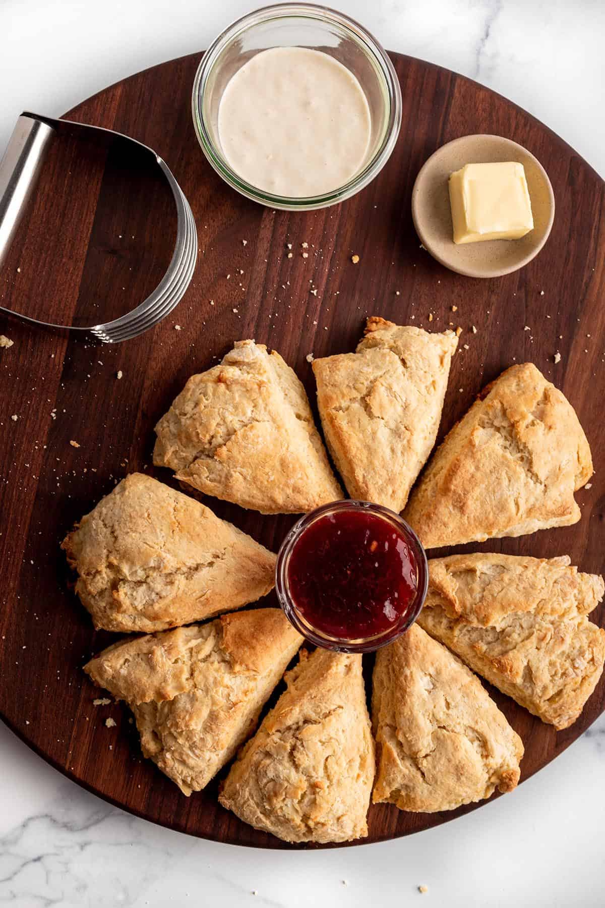 A wooden board with scones, raspberry jam, butter, pastry cutter and a sourdough starter. 