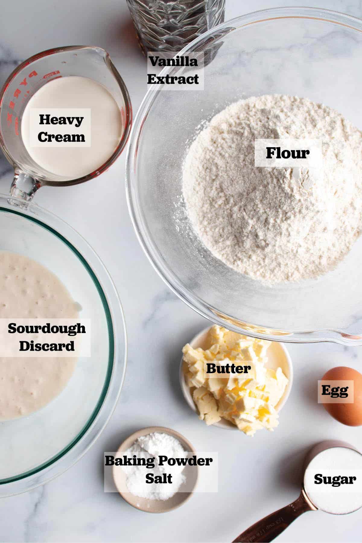 Ingredients for scones laid out in bowls and measuring cups. Vanilla extract, heavy cream, flour, sourdough discard butter, egg, sugar, baking powder and salt. 