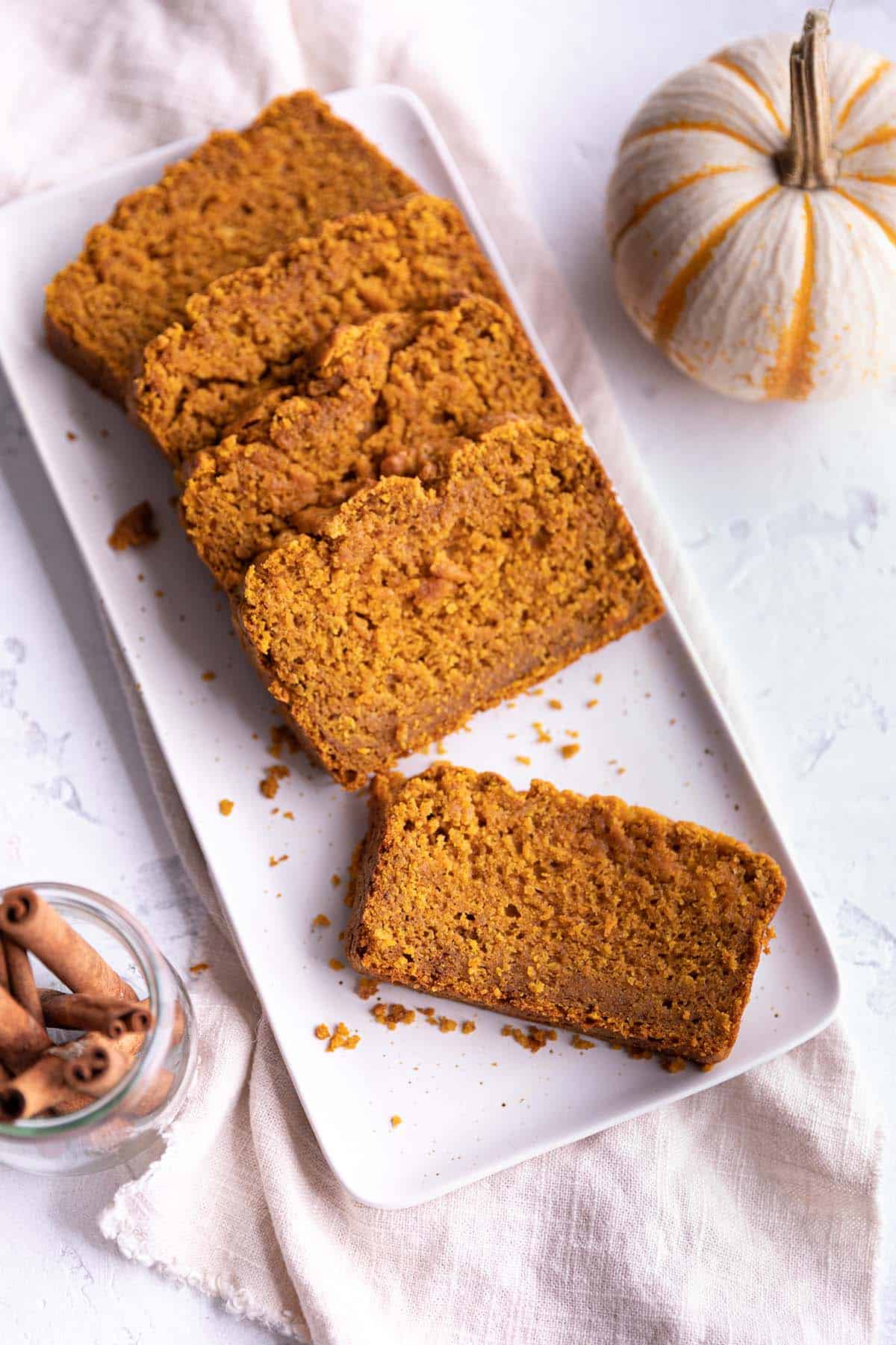 A loaf of pumpkin bread slices on a rectangular plate with a pumpkin and cinnamon sticks. 