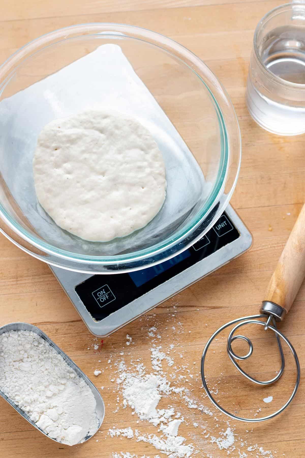 A digital scale with a bowl of sourdough starter, dough whisk, flour and water.