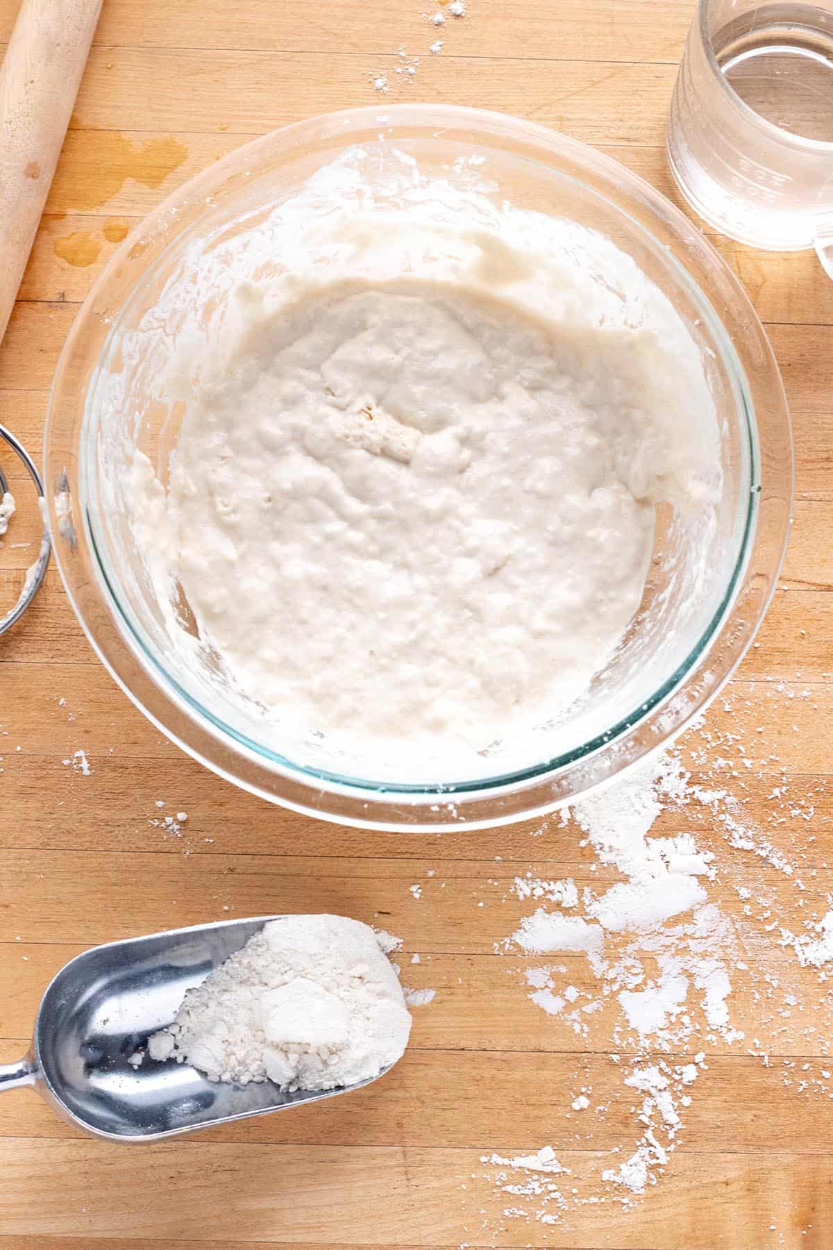 A glass bowl with a sourdough starter, dough whisk, cup of water and scoop of flour.