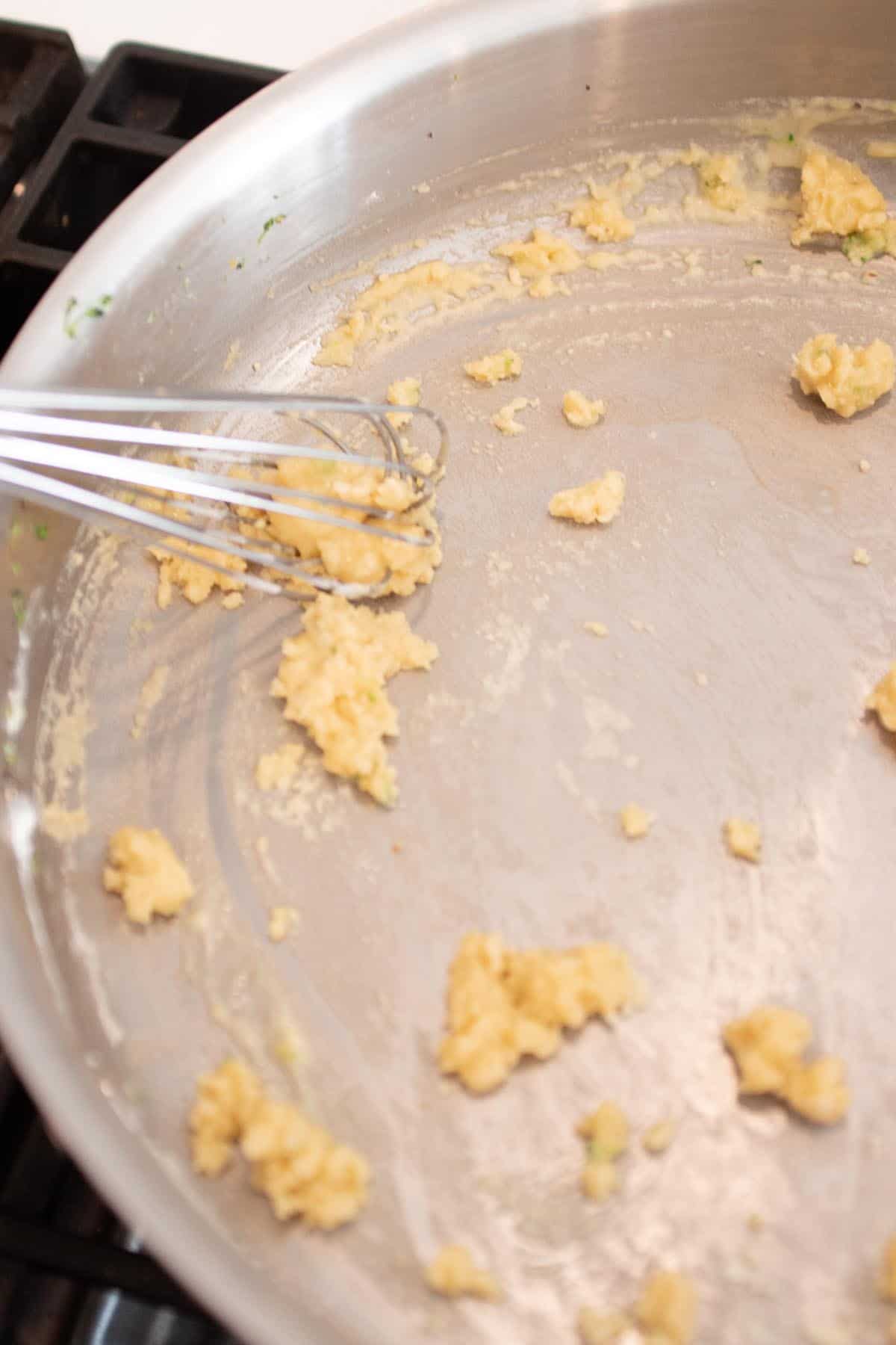 A sauce pan with clumps of flour and butter being mixed with a whisk.