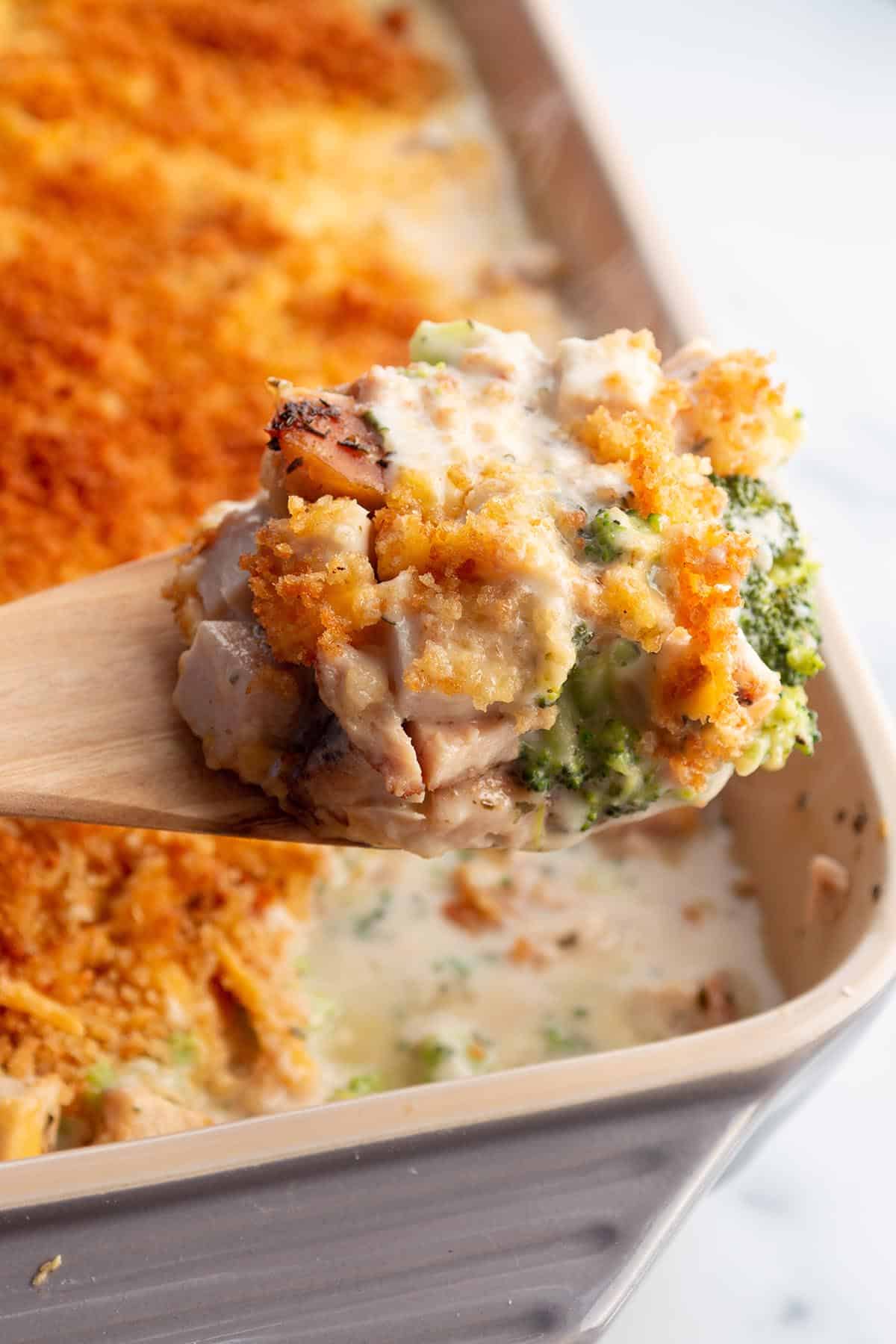 A wooden spoon taking a scoop out of turkey, broccoli and cheese from a baking dish. 