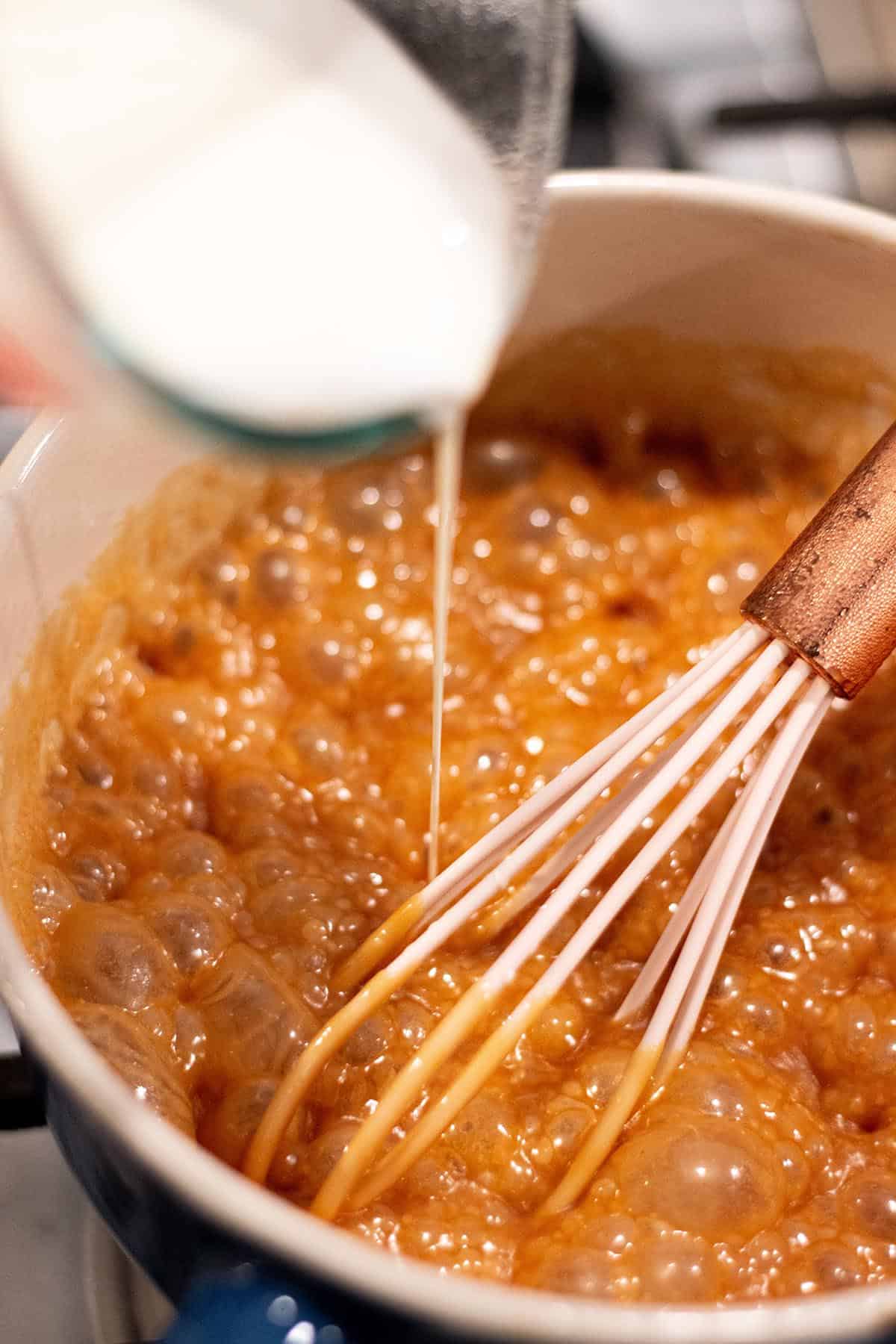 A small sauce pot with a bubbling caramel sauce with cream being whisked in.