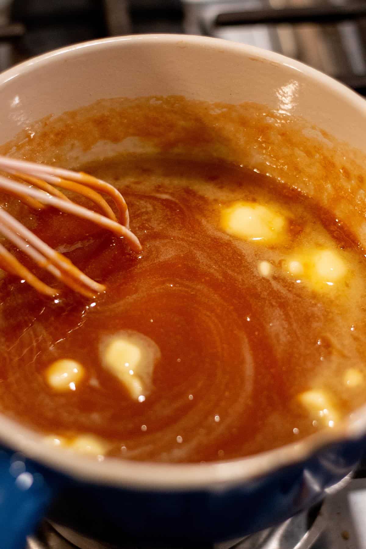 A small sauce pot with caramel sauce and cubes of butter being stirred in.