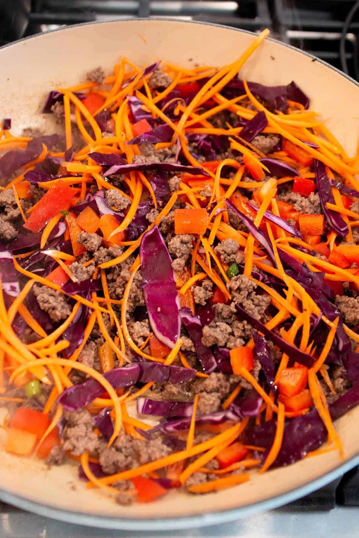 A large braiser with cooked ground beef, julienne carrots, chopped red bell peppers and cabbage. 