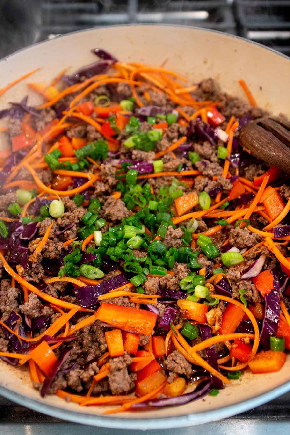 A large braiser with browned ground beef, julienne carrots, chopped cabbage and red bell peppers with green onions on top. 