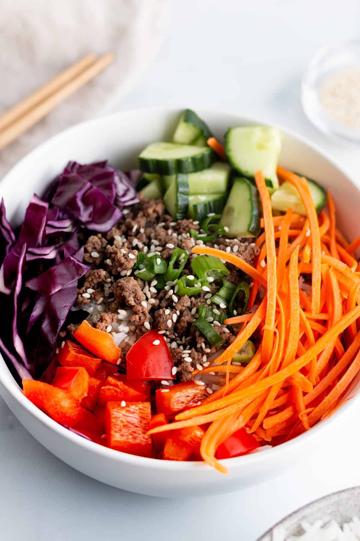 A white bowl with julienne carrots, red bell peppers, cabbage cucumbers and cooked ground beef. 