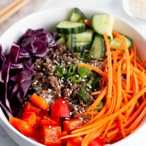 A bowl full of rice, ground beef, julianne carrots, cucumbers, cabbage and red peppers.