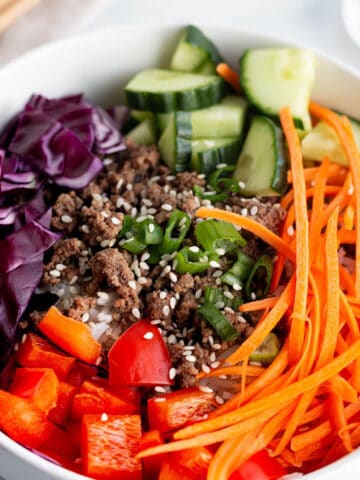 A bowl full of rice, ground beef, julianne carrots, cucumbers, cabbage and red peppers.