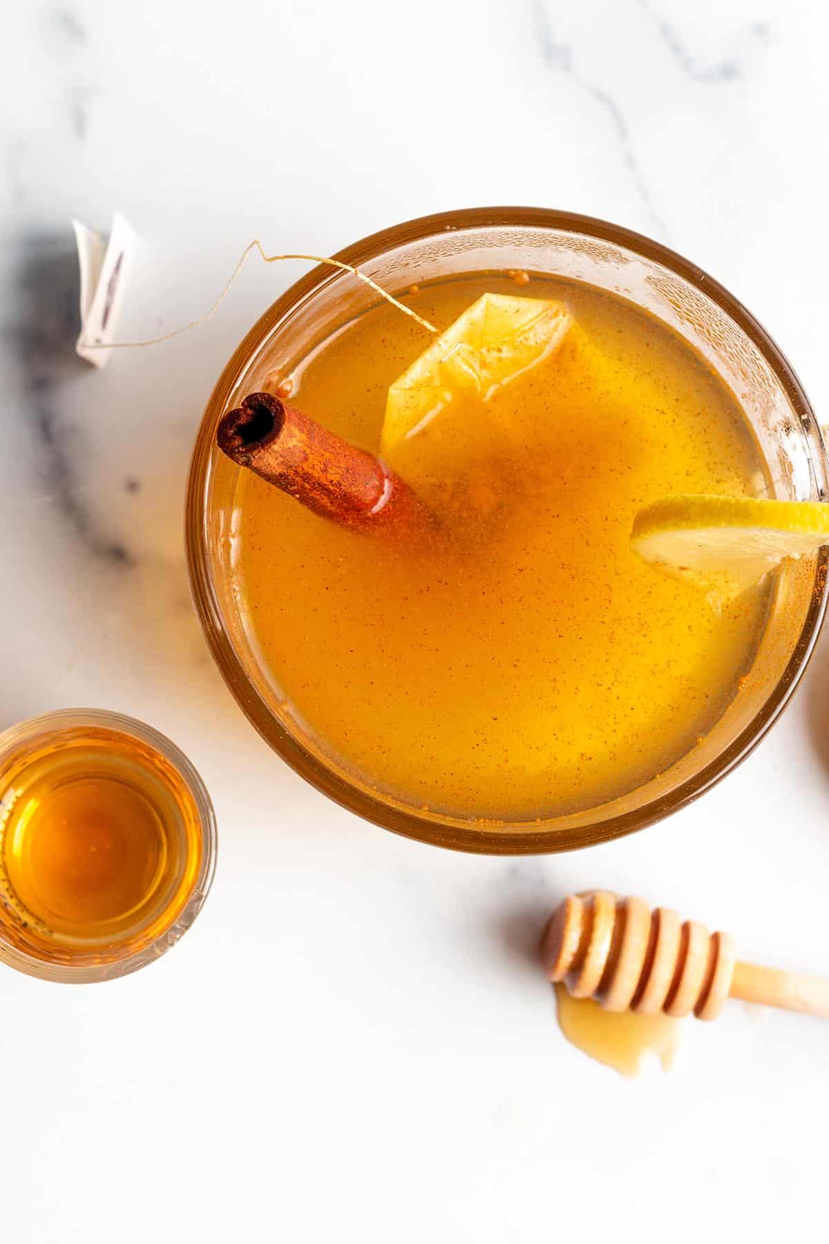 A glass mug with a tea and cinnamon stick, shot of bourbon and honey stick.