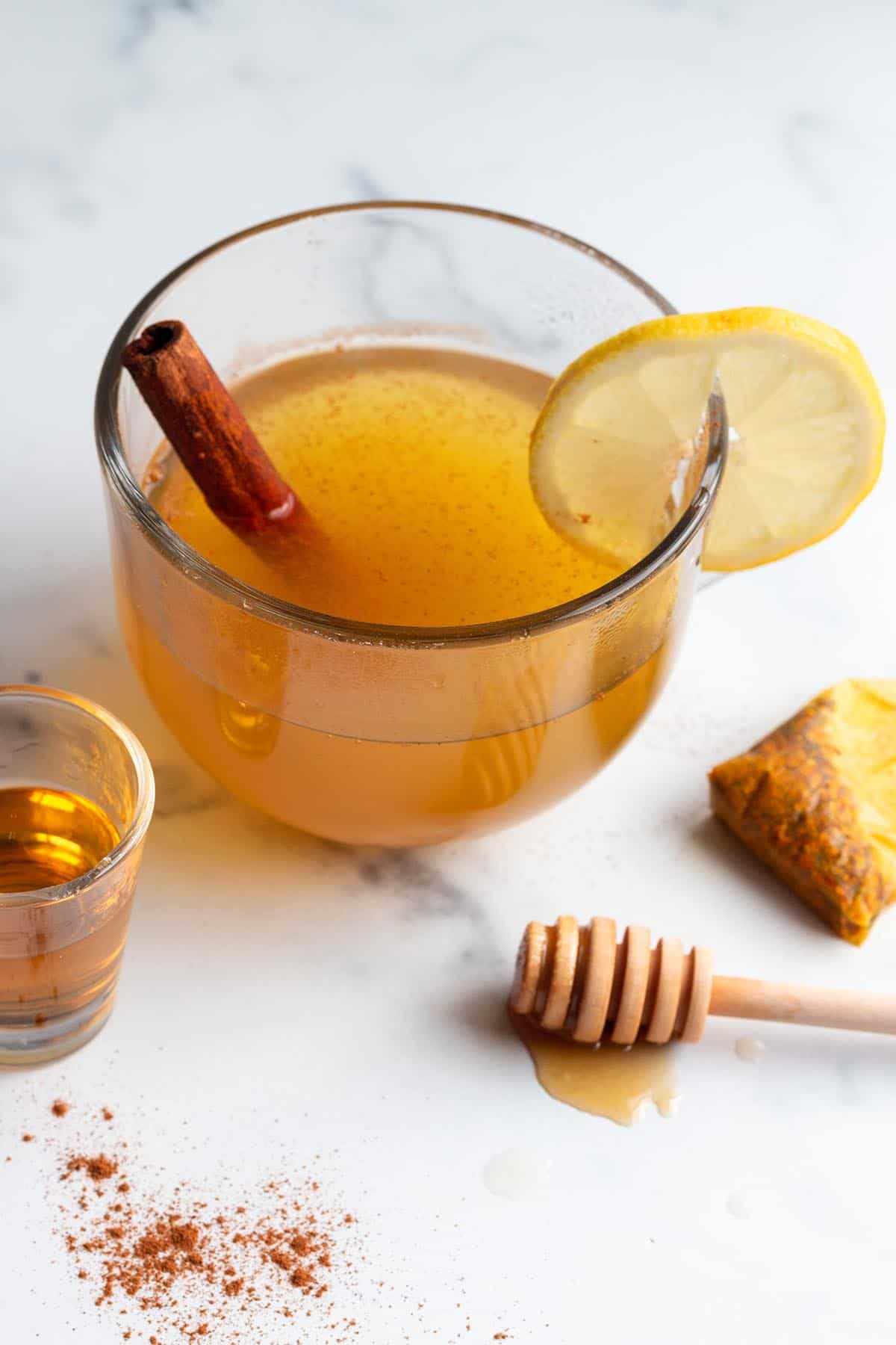 A glass mug with a hot toddy, cinnamon stick, lemon slice, tea bag and honey stick.