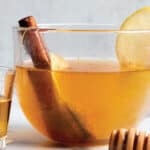 A glass coffee mug with tea steeping in hot water with a honey comb stick and shot of bourbon.