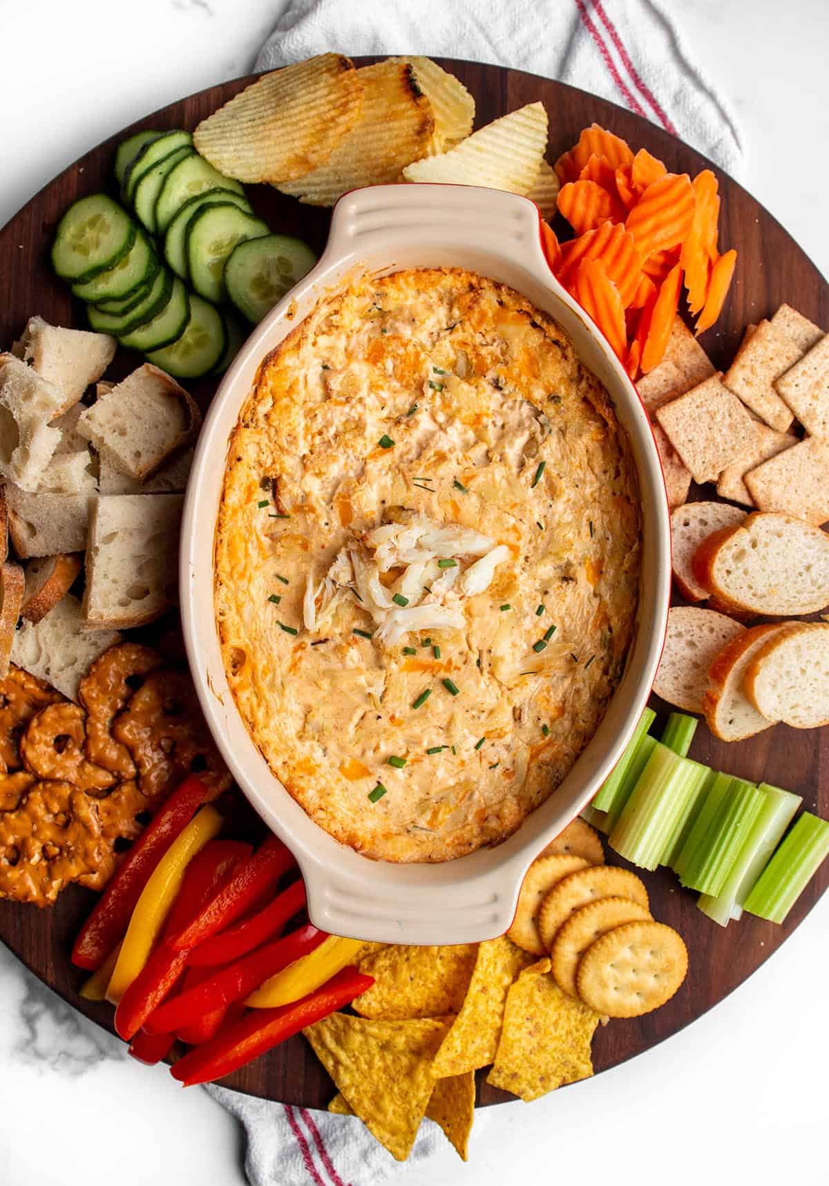 A large circular wood board with a hot crab dip in the middle with fresh produce, bread, chip and crackers around the dish.