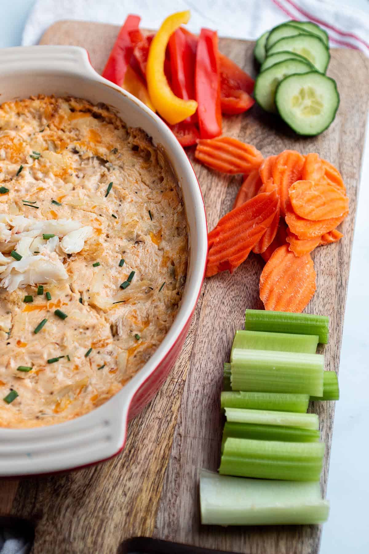 A wooden board with a baking dish full of crab dip with celery, carrots, bell peppers, and cucumber slices. 