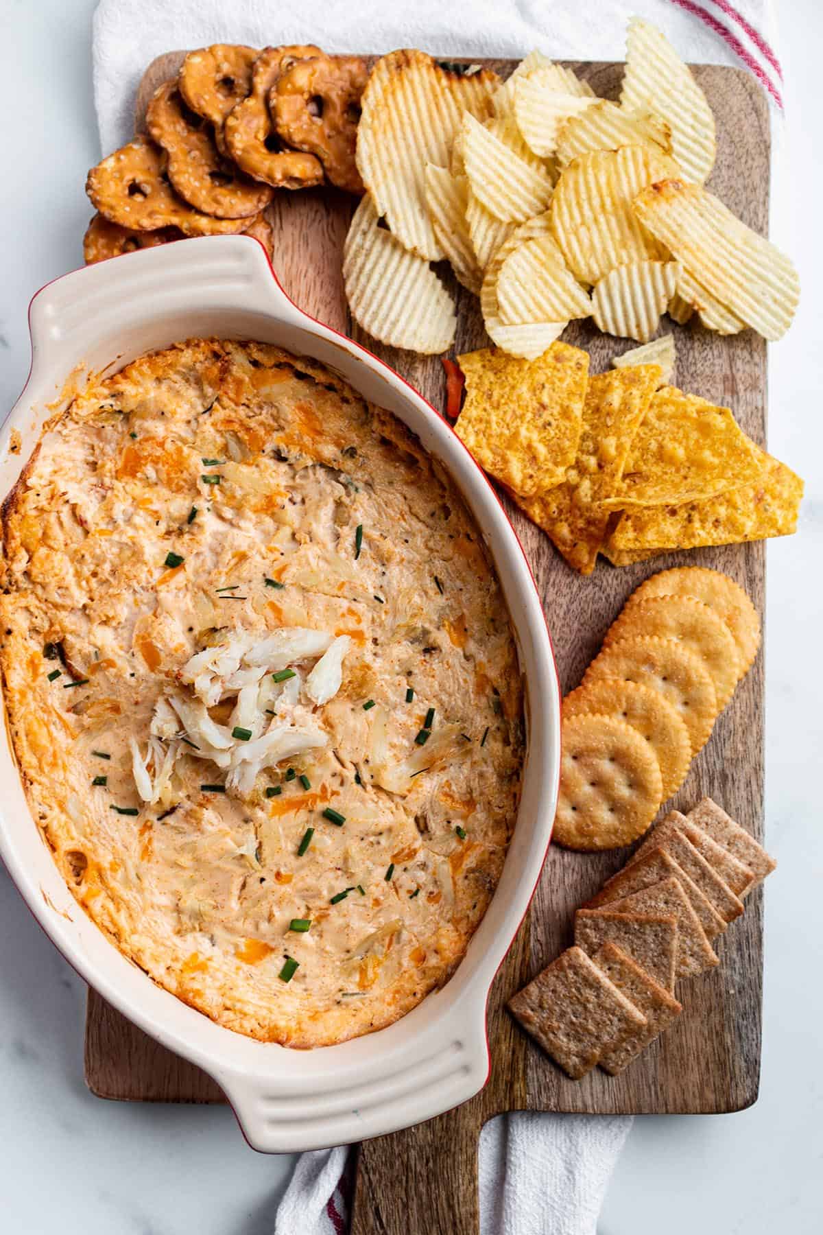 A wooden serving board with a baking dish with crab dip with pretzels, chips and crackers. 