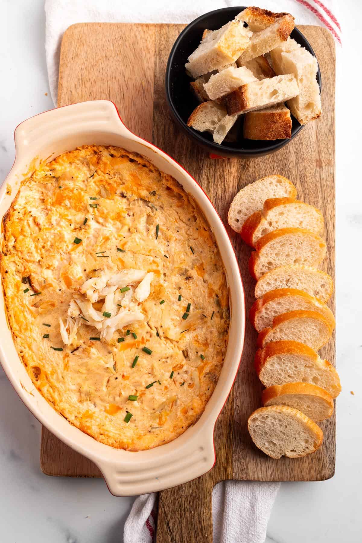 A wooden board with crab dip in a baking dish with sliced baguette and pieces of sourdough. 