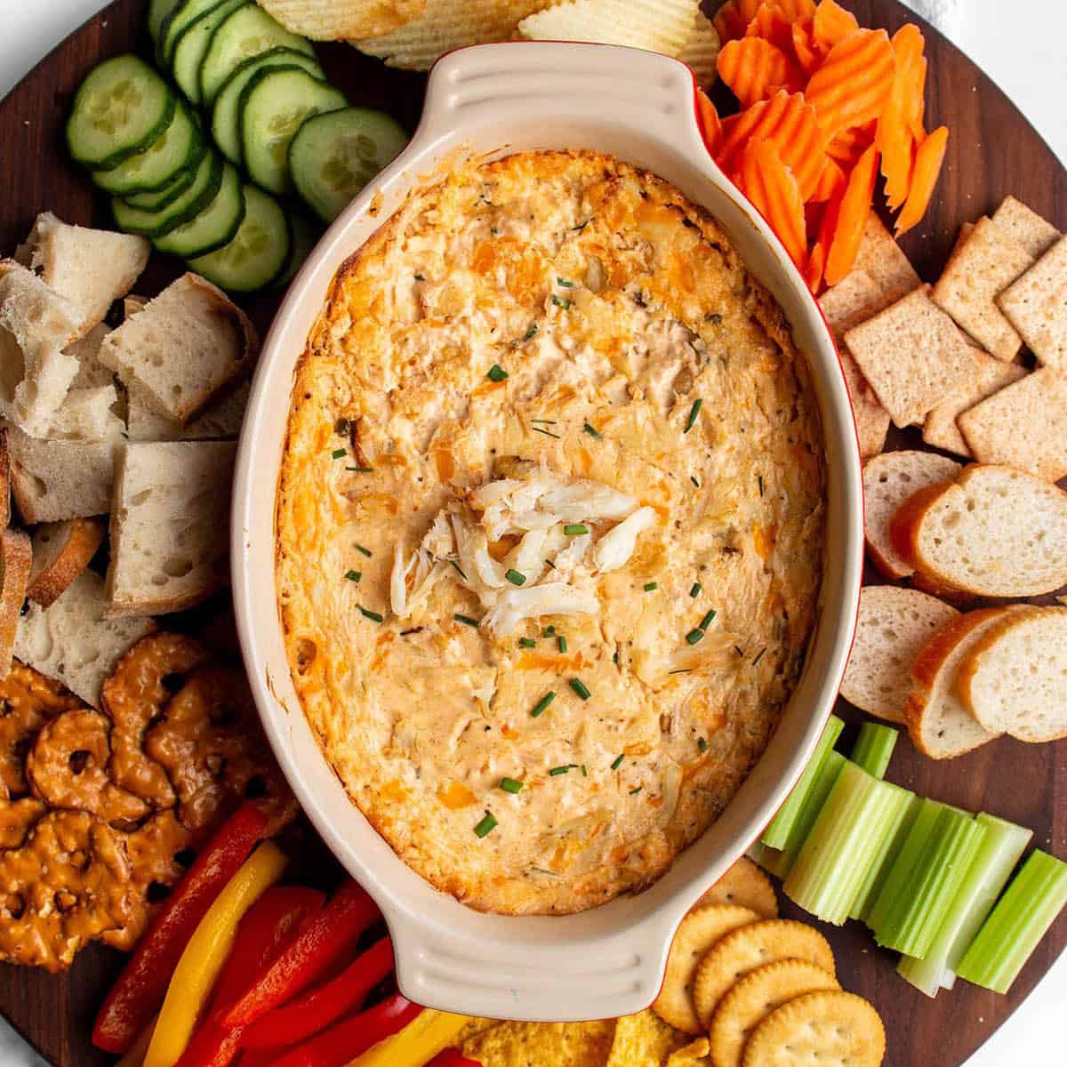 A large circular wood board with a hot crab dip in the middle with fresh produce, bread, chip and crackers around the dish.