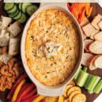 A circle wooden board with a bowl of crab dip surrounded by all kinds of fresh produce, bread, crackers and chips.