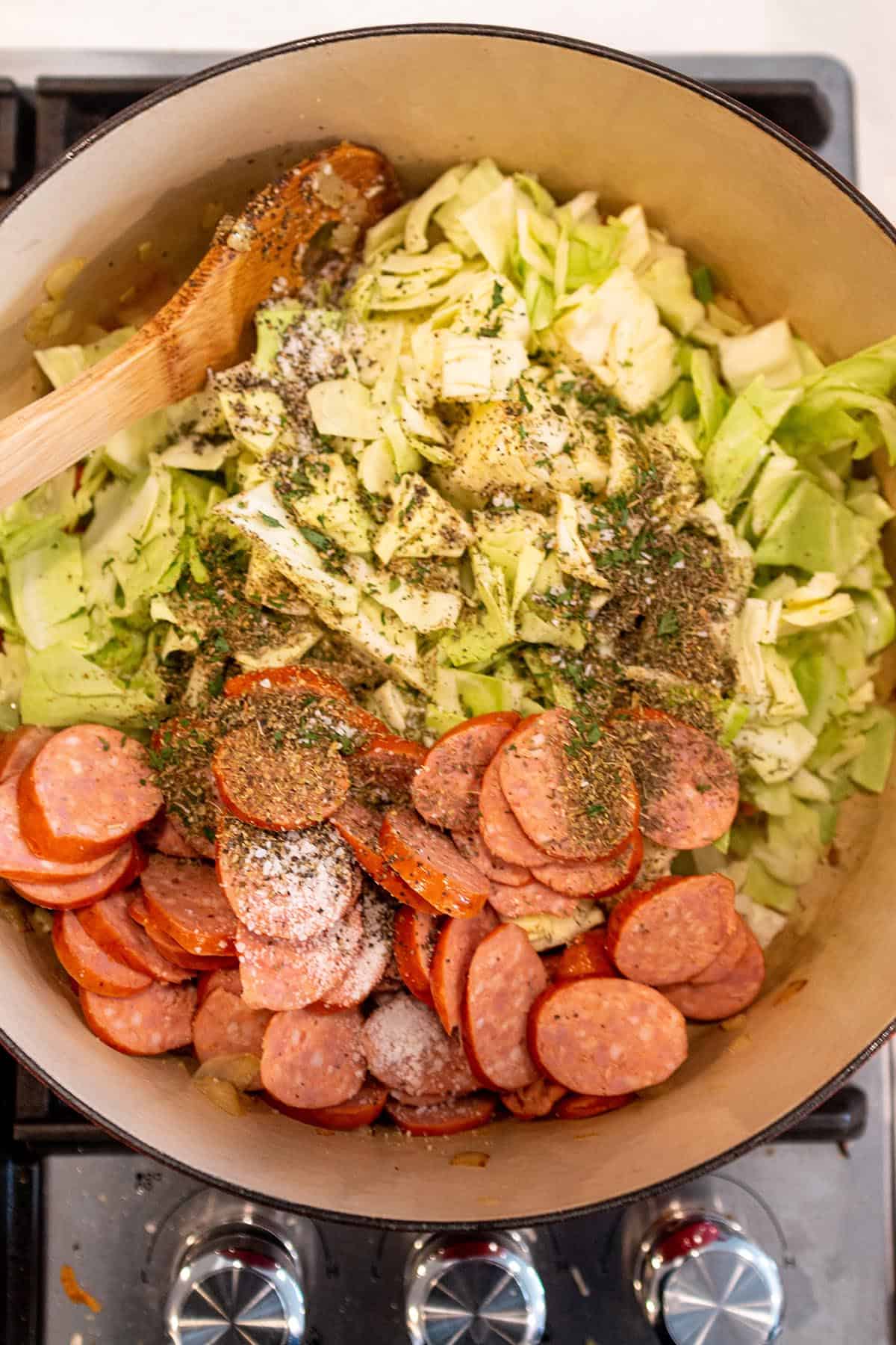 A large pot on the stove with sausage and cabbage being added to a soup.