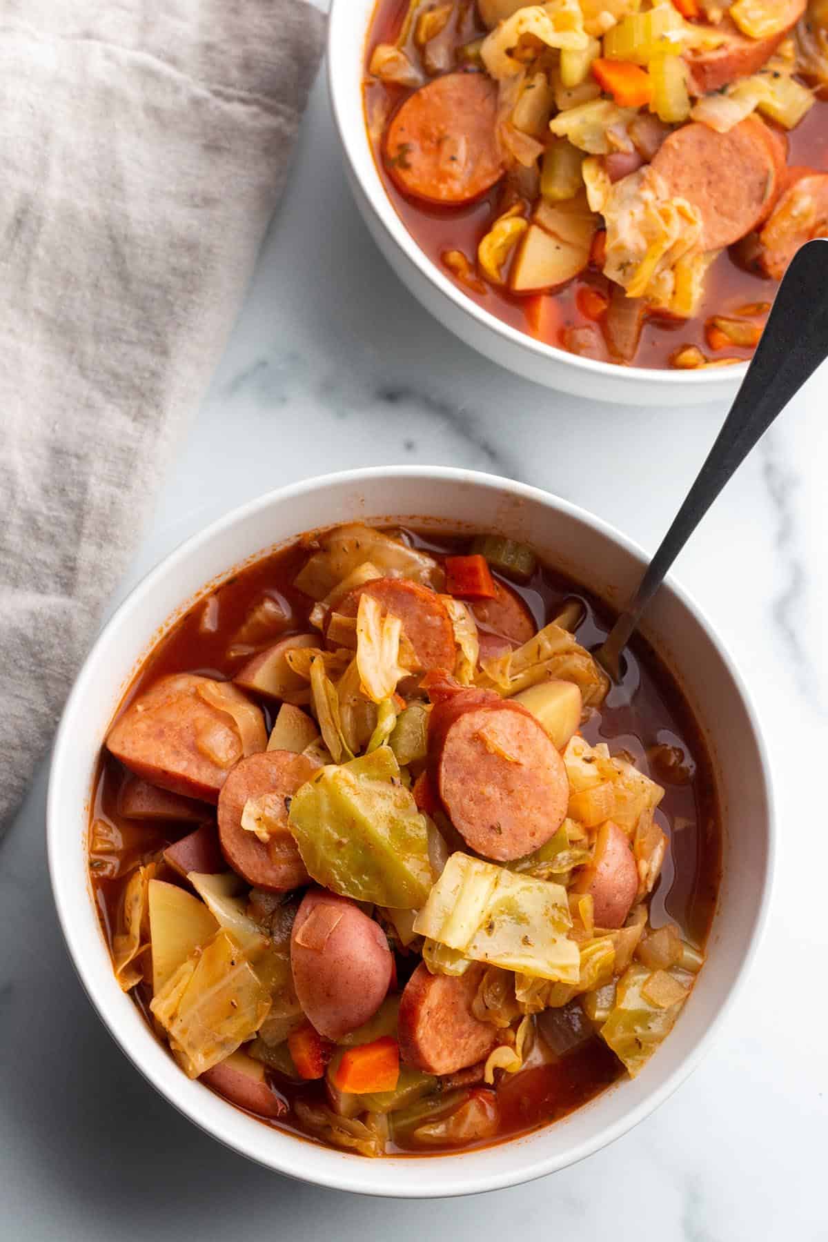 A white bowl full of soup made with sausage links, cabbage leaves and red potatoes with a spoon sticking out.