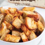 A white bowl with homemade croutons with a loaf of bread in the back.