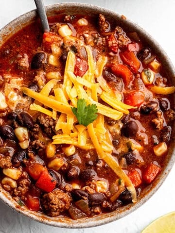 A bowl full of soup made out of ground beef, beans, corn and topped with cheddar cheese.