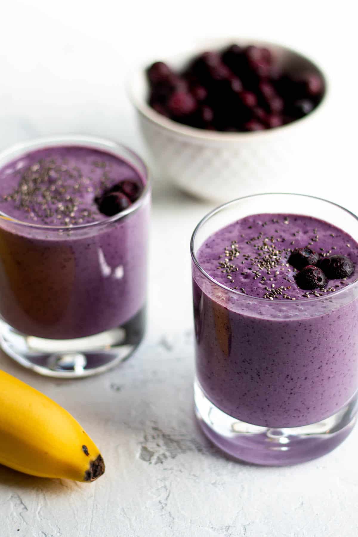 Two small glasses with a purple smoothie, a bowl of blueberries and a banana.
