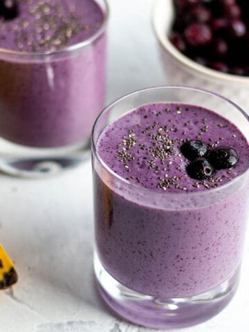 Two small glasses with a purple smoothie topped with blueberries with a bowl of blueberry in the back and a banana.